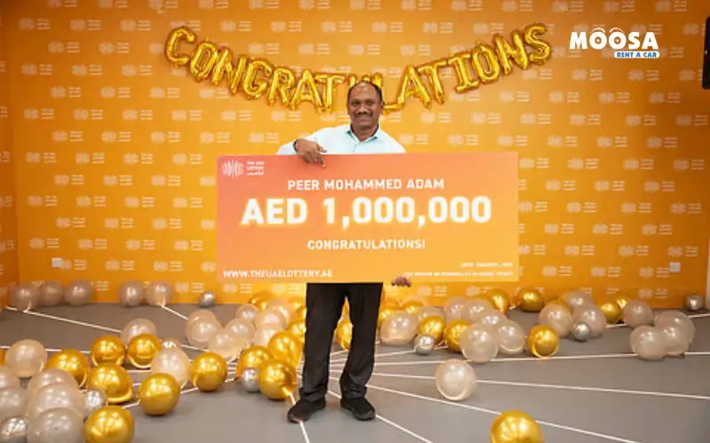 Ein Lotteriegewinner aus den VAE hält einen großen Scheck über 1,000,000 AED in einer festlichen Umgebung mit goldenen und silbernen Luftballons und einem „Herzlichen Glückwunsch“-Banner.