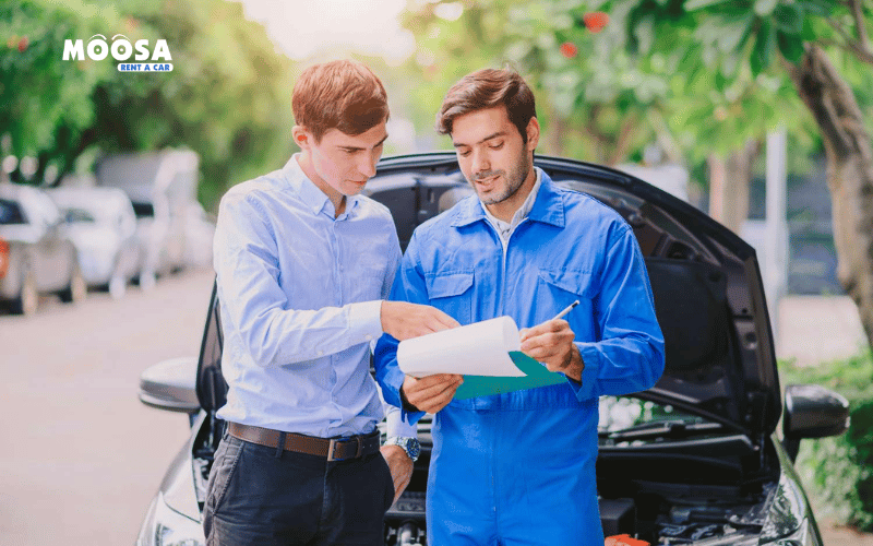 Mechanic in blue coveralls discussing car insurance details with a client near an open hood, set in a green, outdoor environment.