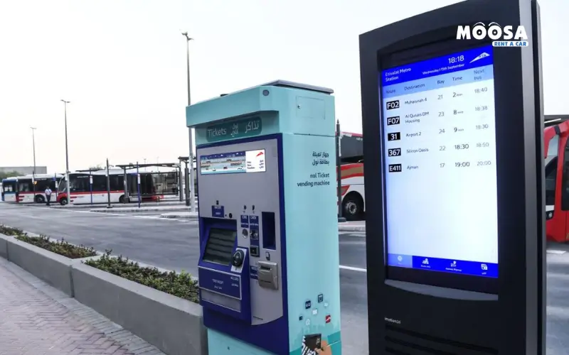 Ticket vending machine and digital schedule display at a bus station, with buses parked in the background and clear signage.