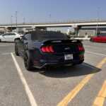A sleek black convertible Mustang GT parked in a sunlit lot, with a white sedan and a red car in the background, under a clear sky.