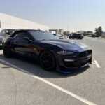 A sleek black convertible Mustang GT with blue accents parked in a lot, surrounded by other cars under a clear sky.