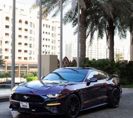 A sleek purple Ford Mustang GT parked in a modern urban setting with palm trees and skyscrapers in the background.