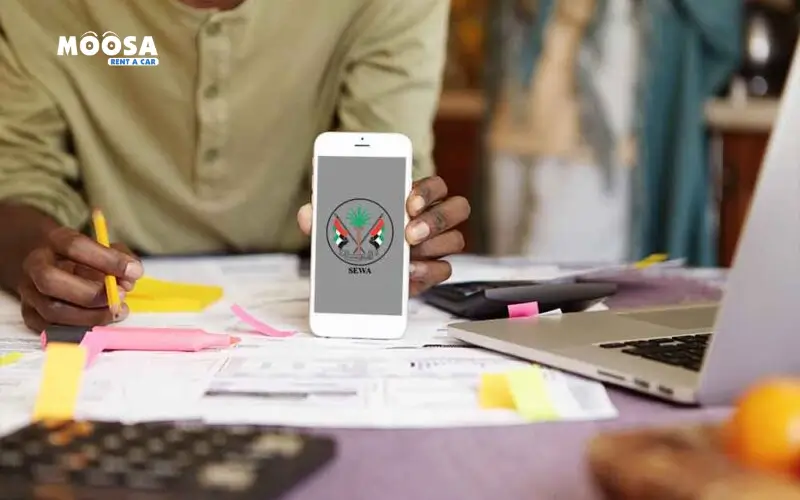 A person holds a smartphone displaying a sewa mobile app logo on a cluttered desk with papers, sticky notes, and a laptop in the background.