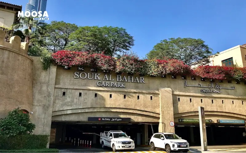 Entrance to Souk Al Bahar car park in Dubai, featuring lush greenery and a clear blue sky. Two cars parked near the entrance.