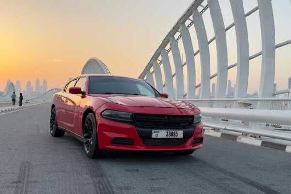 A red Dodge Charger parked on a modern bridge at sunrise, with city skyscrapers in the background and two people in silhouette.